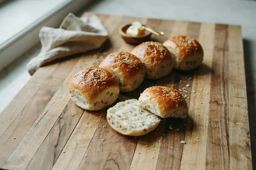 Six whole grain hamburger buns in a row with sesame-topped domes, one sliced open showing a soft sturdy interior.