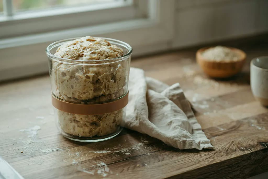 Fresh milled flour dough risen in a straight-sided glass container, bubbles visible through the side, with a rubber band marking the original level.