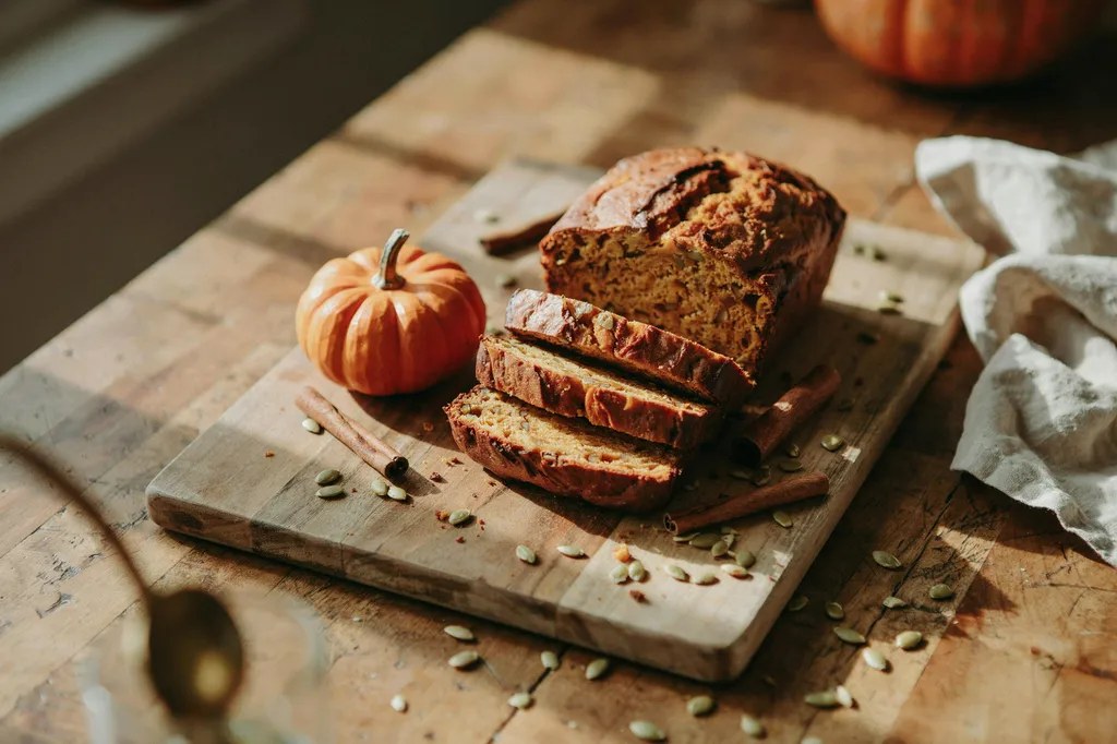 A pumpkin bread loaf with a cracked domed top, slices showing a moist dense orange crumb, with pepitas scattered around.