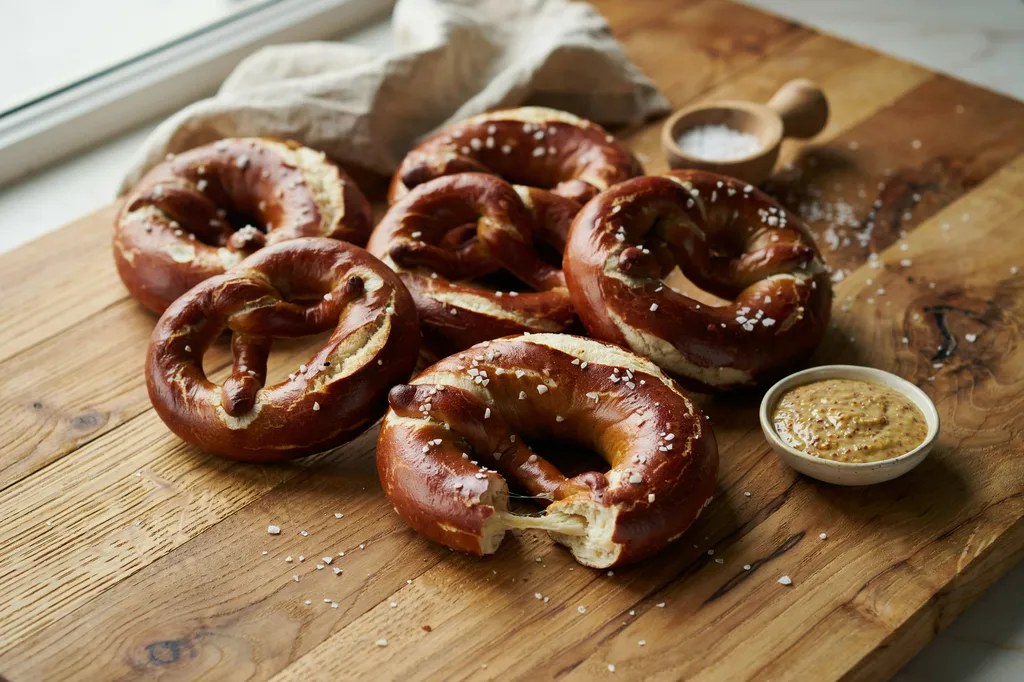 Five large knotted soft pretzels with deep mahogany crusts and coarse salt, one torn open showing the soft interior.