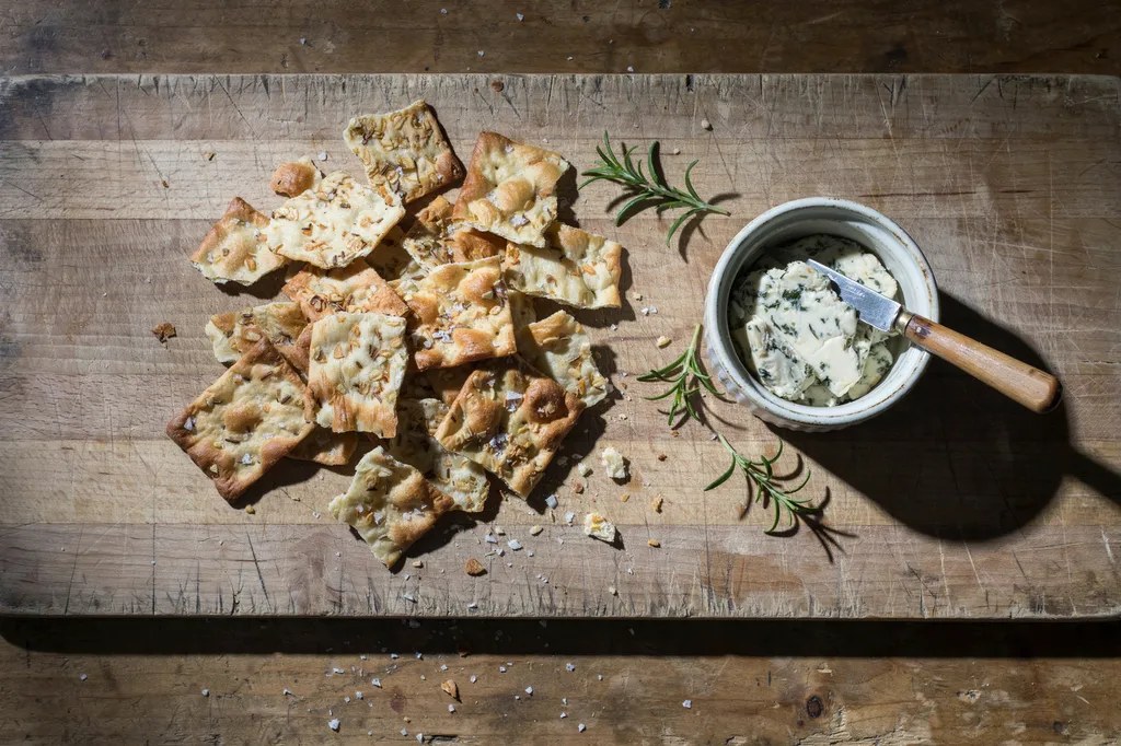 A pile of thin, irregular whole grain sourdough discard crackers scattered with coarse sea salt, beside a ramekin of herbed cheese.