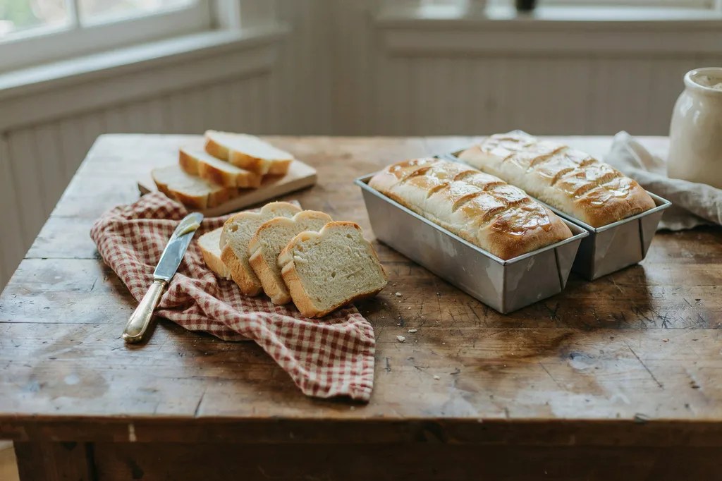 Two soft golden Amish-style sandwich loaves on a farmhouse table, one turned out and sliced showing a fine soft crumb.