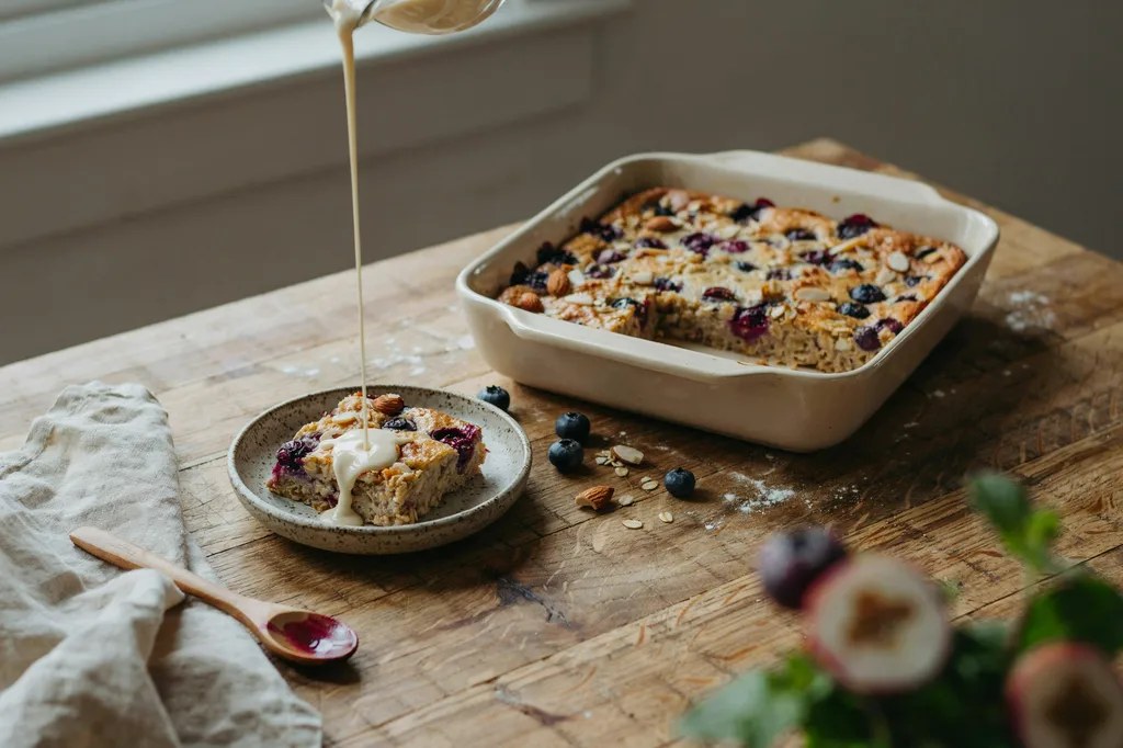 A dish of baked oatmeal studded with blueberries and almonds, one square lifted into a bowl with cream poured over.