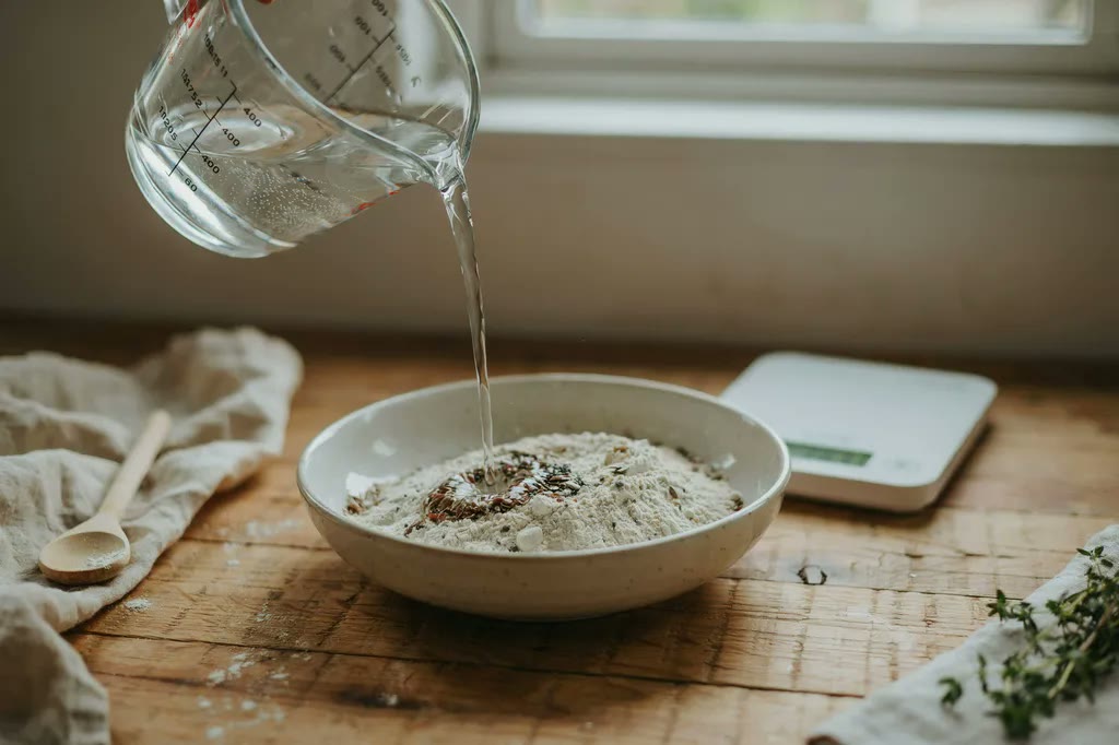 Water being poured from a glass jug into a bowl of speckled fresh milled flour, beside a digital kitchen scale.