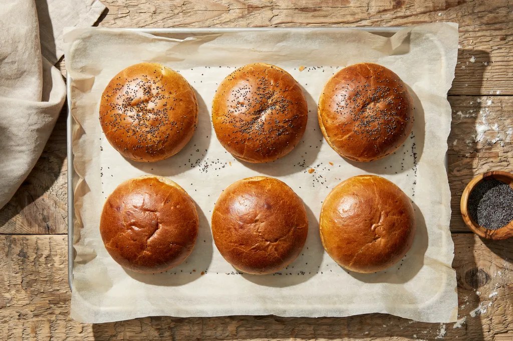 Overhead view of eight round whole grain burger buns on a parchment-lined tray, half topped with poppy seeds.
