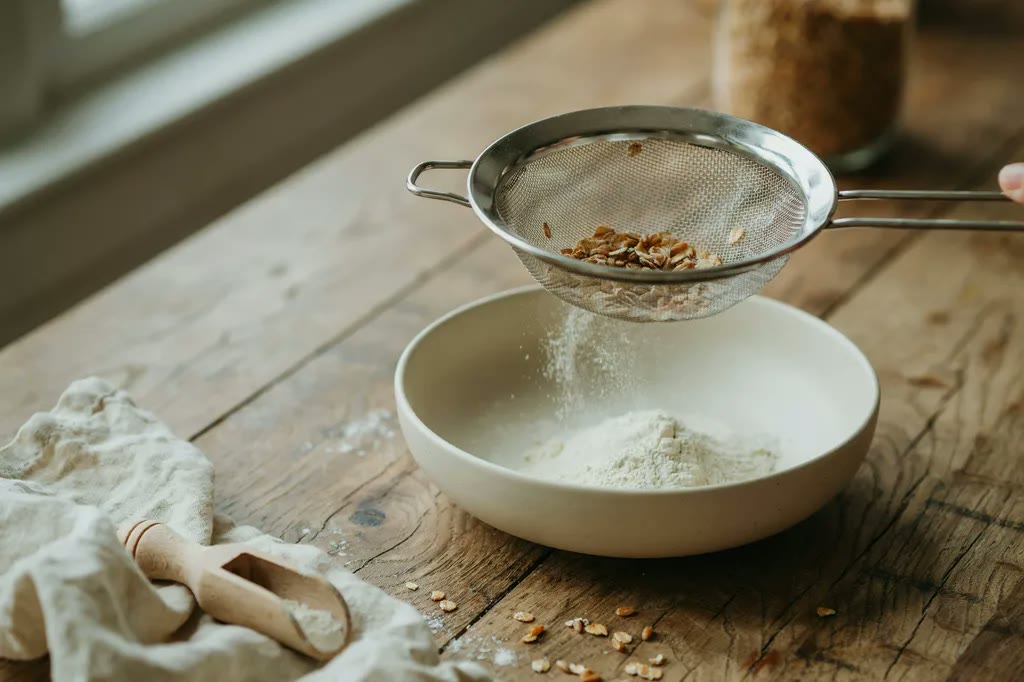Fine flour falling through a mesh sieve into a bowl while coarse bran flakes stay behind in the mesh.