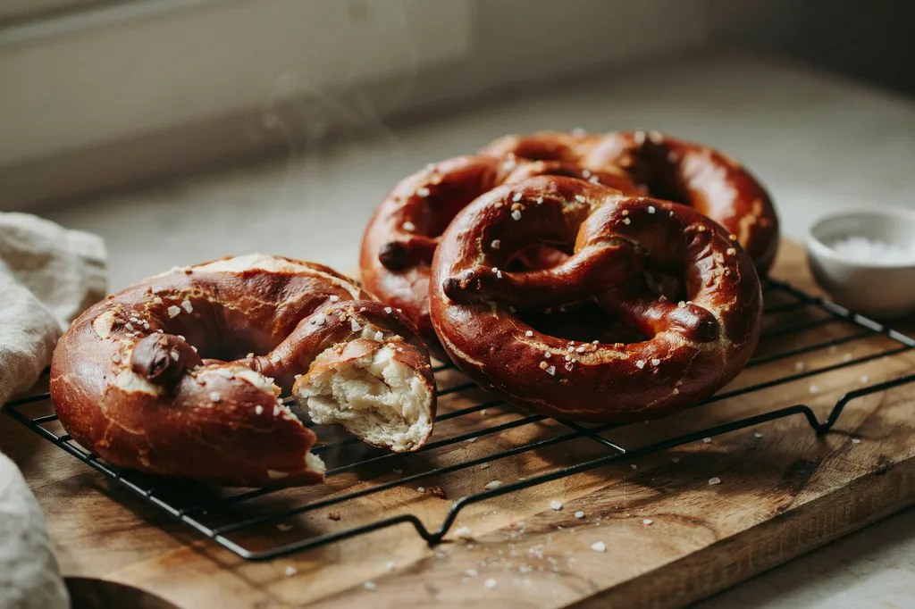 Four large knotted soft pretzels on a wire rack with glossy mahogany crusts and coarse salt, one torn apart.