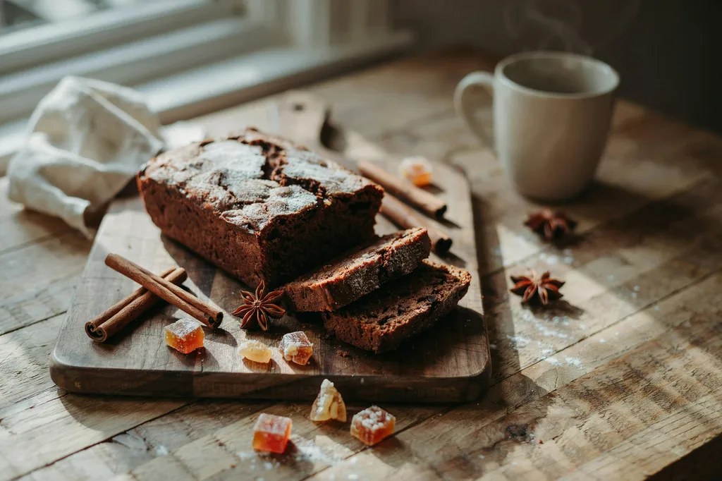 A dark molasses gingerbread loaf dusted with powdered sugar, slices showing a very dark moist crumb, with whole spices around it.
