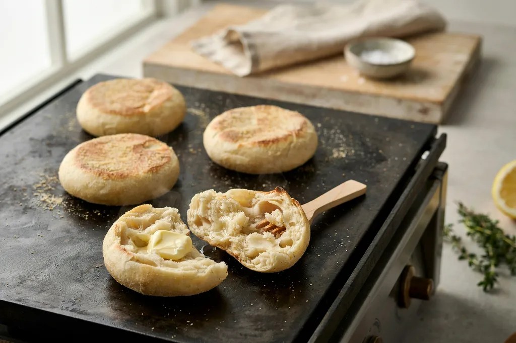 Sourdough English muffins on a griddle, one fork-split showing dramatically craggy nooks with butter melting into them.
