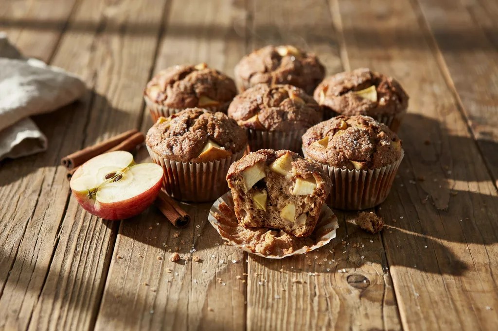 Apple cinnamon muffins with cinnamon-sugar crusted tops, one broken open showing chunks of baked apple in a spiced crumb.