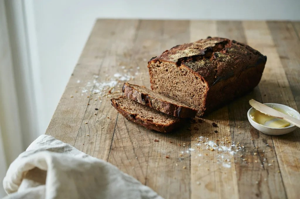 A dark, dense 100% rye pan loaf dusted with rye flour, two slices cut showing a tight moist dark crumb.