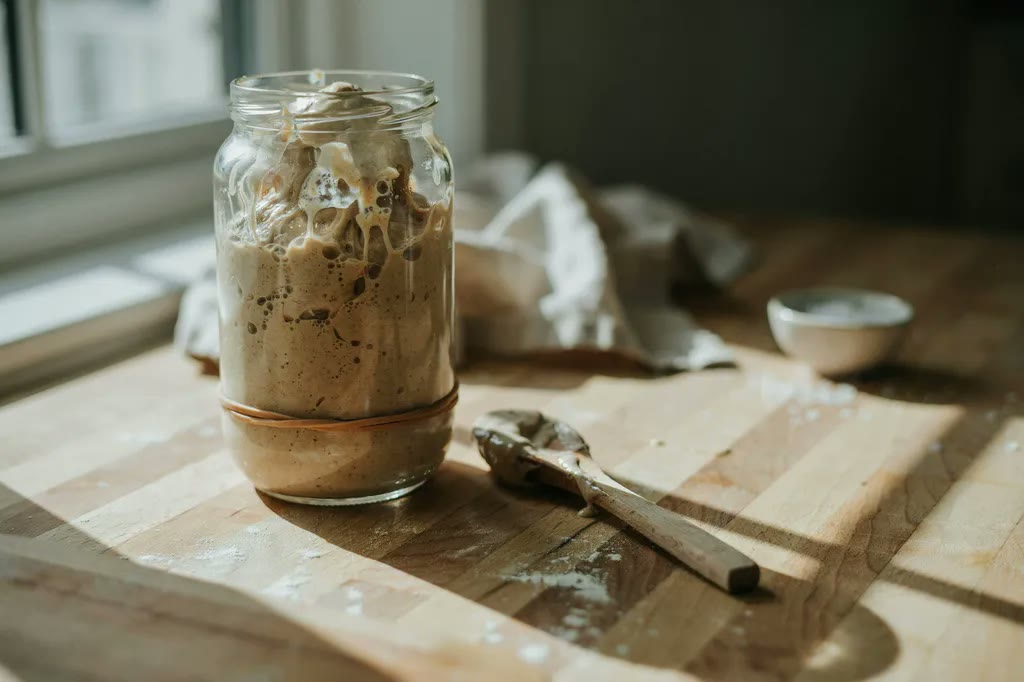 An active whole grain sourdough starter risen and bubbling in a glass jar, with a rubber band marking its starting level.