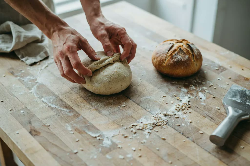 Hands shaping whole grain dough into a taut oval batard on a floured bench, with a shaped round boule beside it.