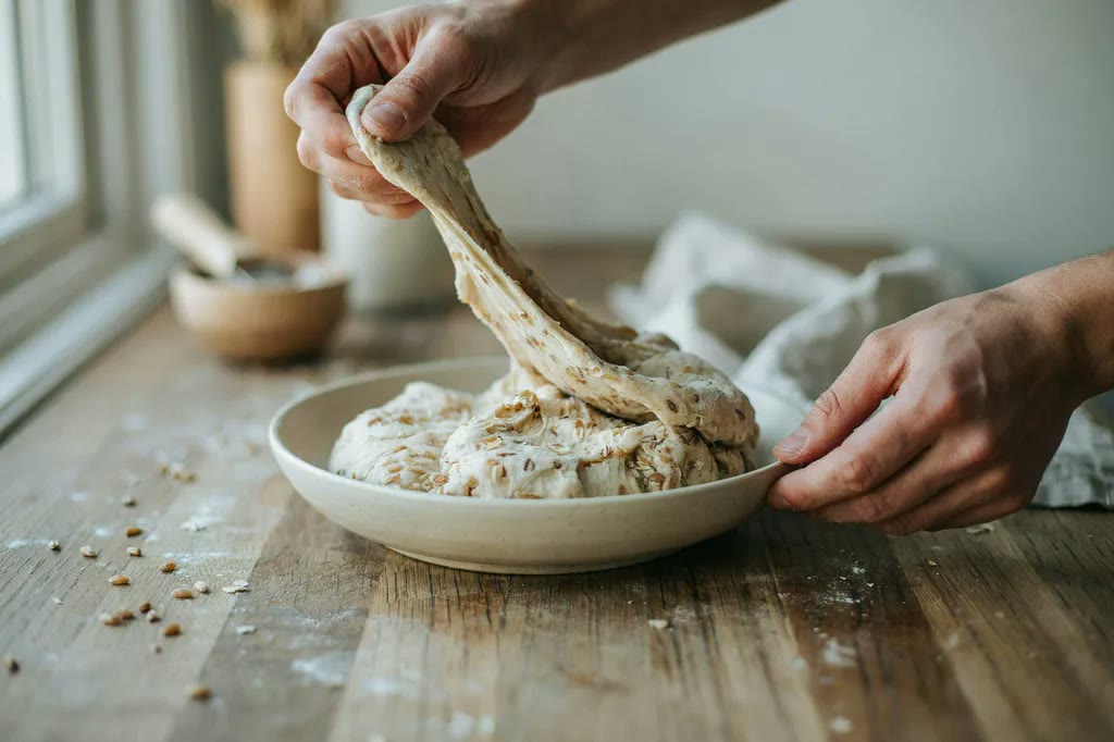 A hand lifting the edge of slack whole grain dough into a long stretch before folding it back over the mass.