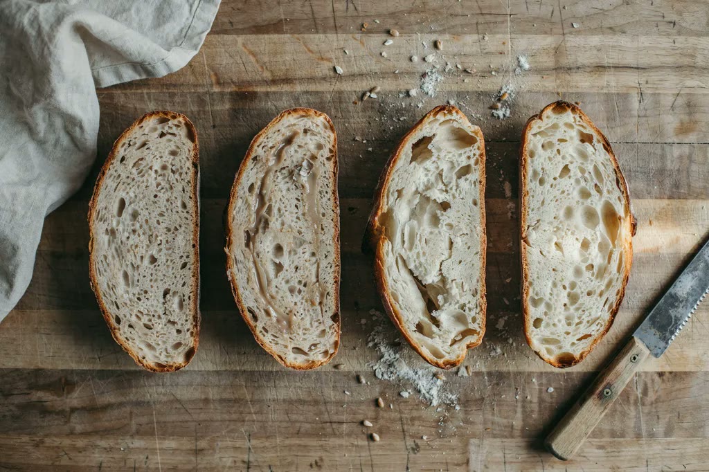 Four halved whole grain loaves side by side showing different crumb faults: dense, gummy, crumbly and open.