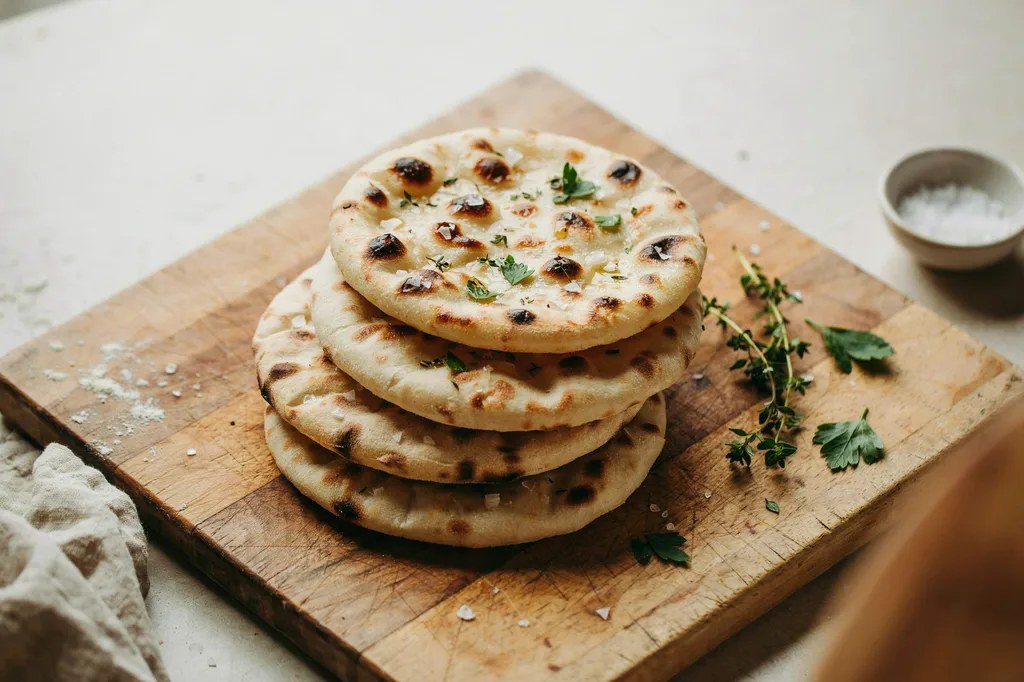 Soft whole grain flatbreads stacked on a board, blistered with dark char spots and scattered with herbs.