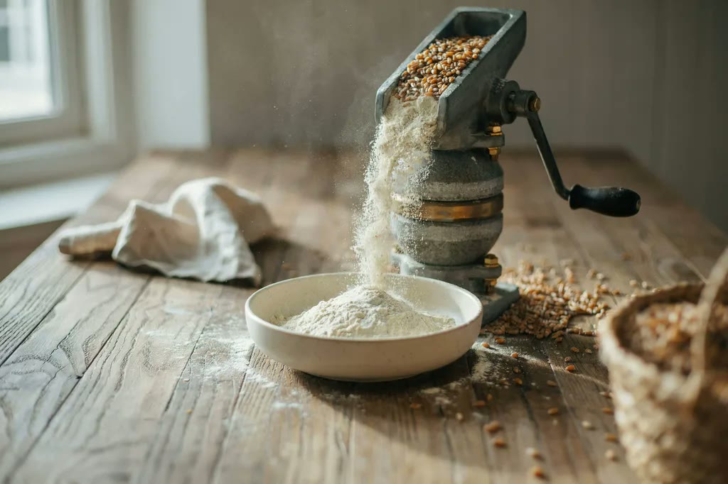 A countertop stone grain mill in use, wheat berries in the hopper and fresh flour falling into a bowl below.