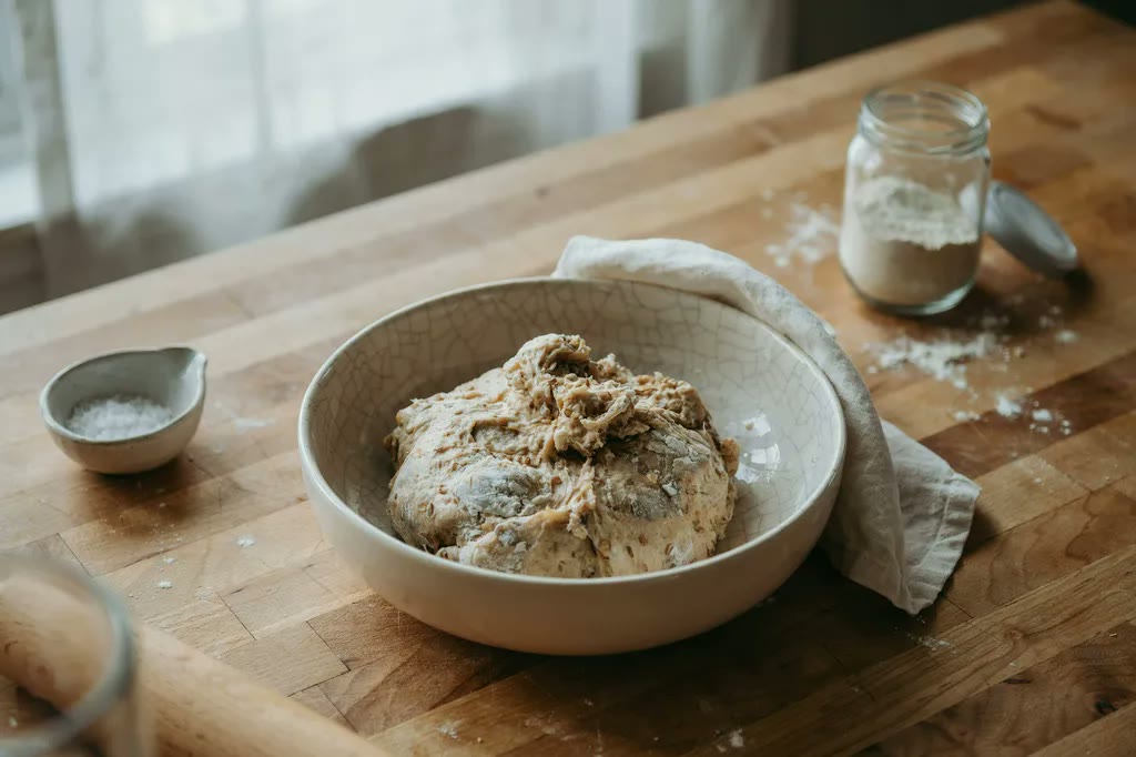 A rough, shaggy mass of freshly mixed whole grain dough resting in a ceramic bowl, flour and water barely combined, before kneading begins.
