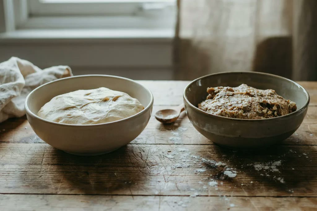 Two bowls side by side, one holding smooth pale white-flour dough and one holding darker, bran-flecked fresh milled dough.