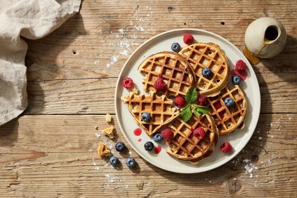Four thin round whole wheat waffles overlapping on a platter, scattered with raspberries and blueberries.